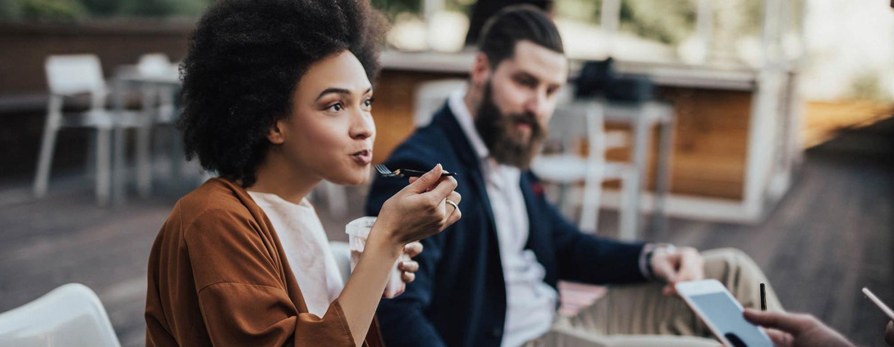 woman sits and eats outside while someone holding a cell talks to her