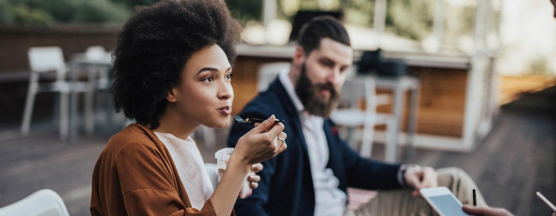 woman sits and eats outside while someone holding a cell talks to her